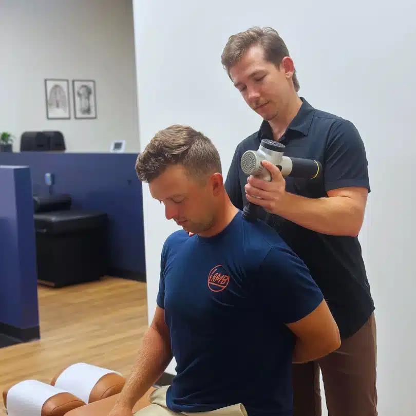 Chiropractor using a percussion massage gun for muscle therapy on patient's shoulders in Fort Worth.
